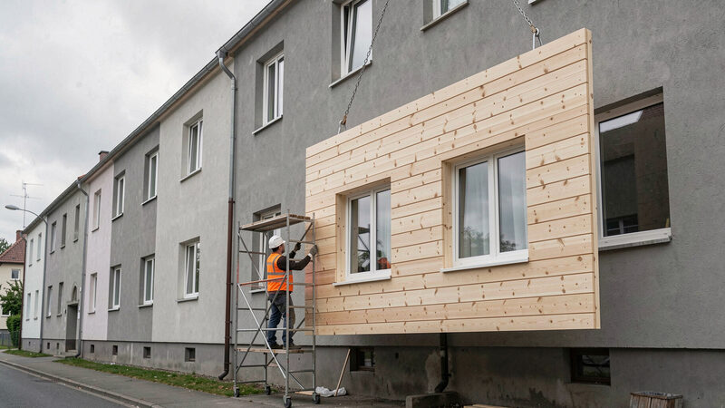 Ein geschosshohes Holzfassadenelement mit zwei eingebauten Fenstern hängt an Ketten vor der Putzfassade eines Mehrfamilienhauses, ein Monteur auf dem Rollgerüst führt es